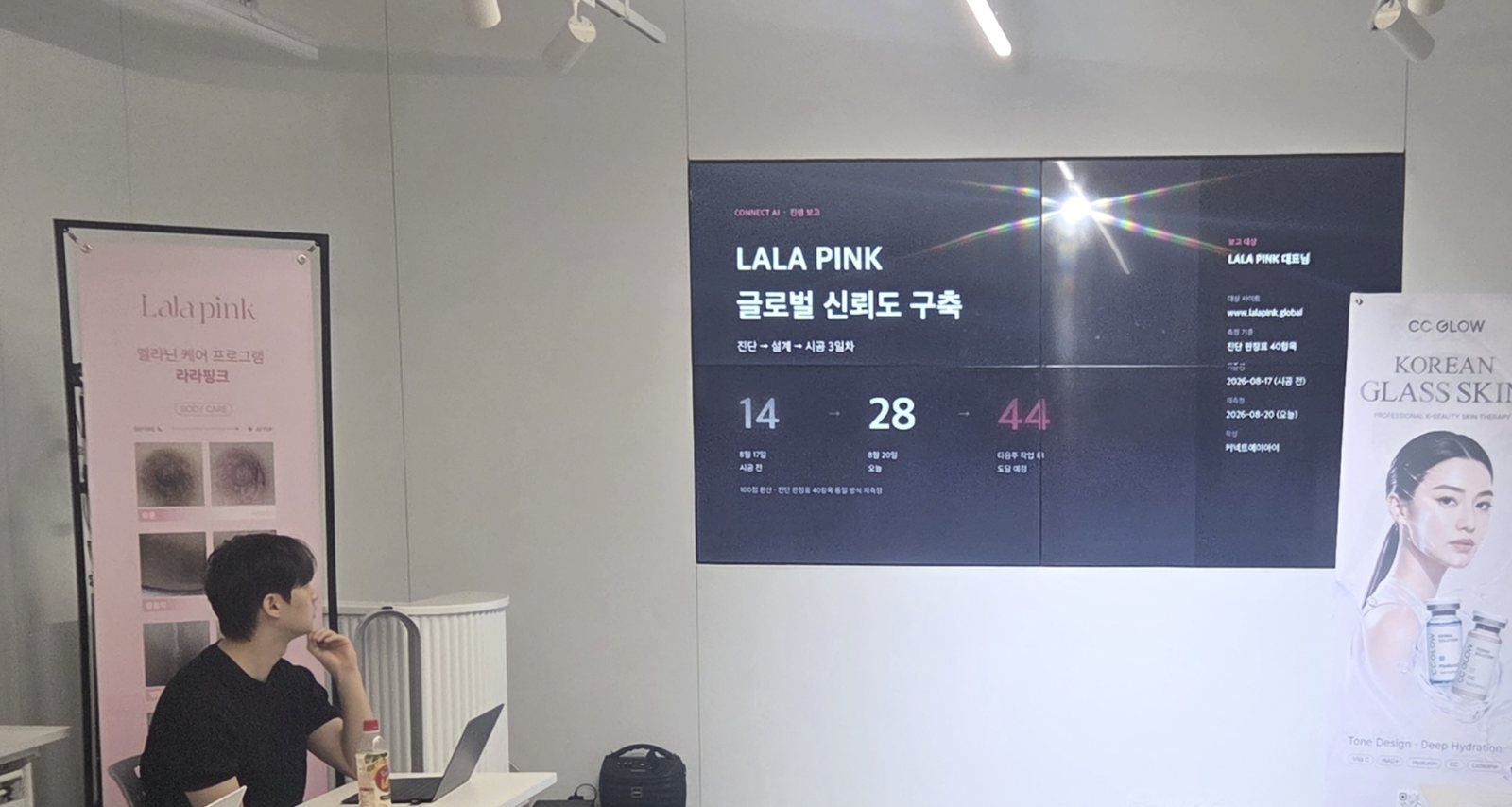 라라핑크 사무실 벽 화면에 「LALA PINK 글로벌 신뢰도 구축 — 진단 → 설계 → 시공 3일차」 보고가 띄워져 있고, 정한영 전문가가 이를 보고 있다